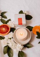 A lit white soy candle in a ribbed holder rests on a plate with white flowers, green leaves, blood orange and orange slices. In the background is a boxed Candle Refill - Amalfi by Mimi & August, releasing a Mediterranean scent.