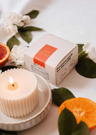 The Candle Refill - Amalfi by Mimi & August, crafted from natural soy wax, sits in a dish beside its box and is accented with green leaves, white flowers, and sliced citrus for a fresh Mediterranean vibe.