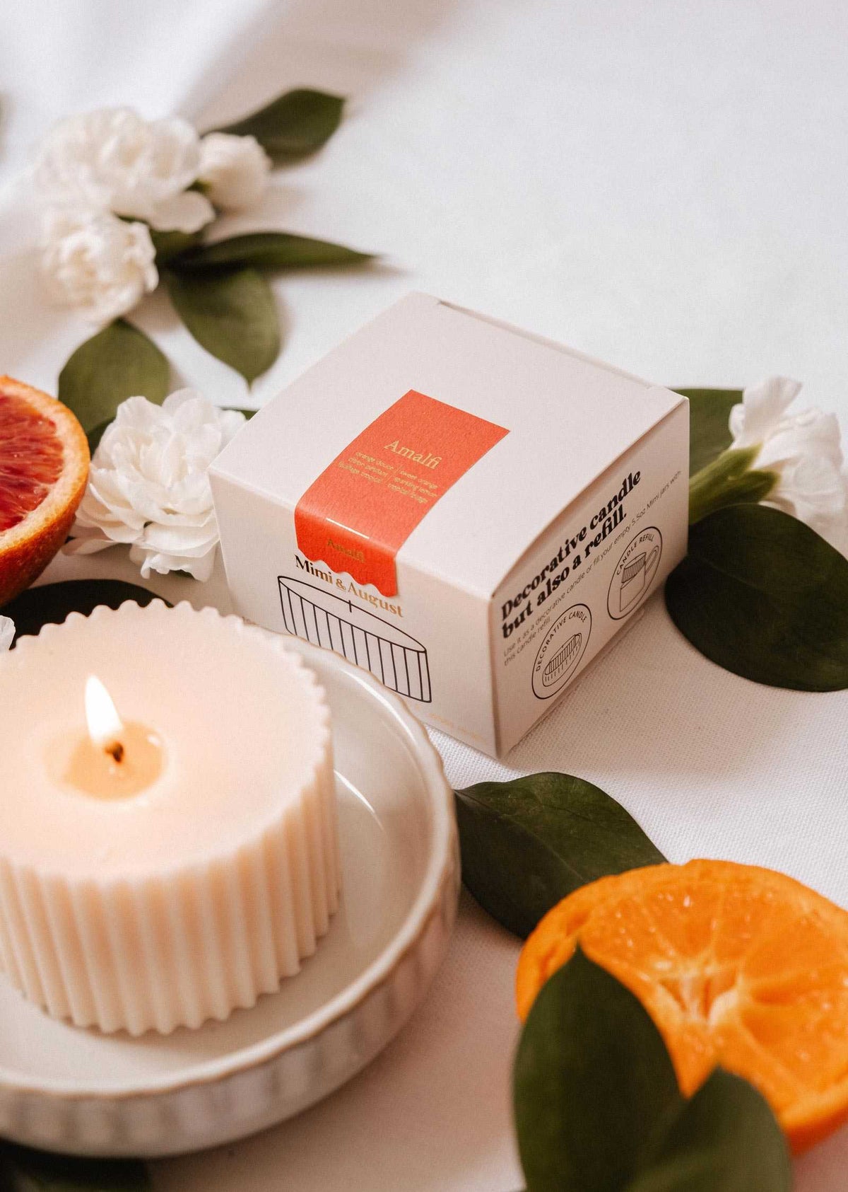 The Candle Refill - Amalfi by Mimi & August, crafted from natural soy wax, sits in a dish beside its box and is accented with green leaves, white flowers, and sliced citrus for a fresh Mediterranean vibe.
