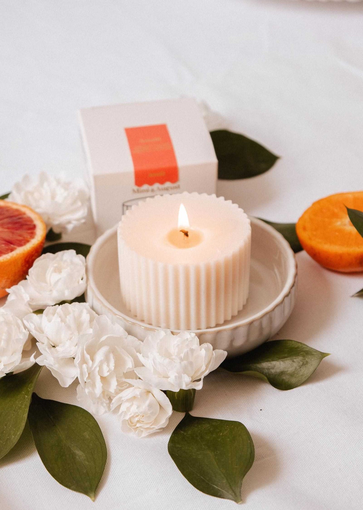 The Candle Refill - Amalfi by Mimi & August, a white ribbed soy wax candle, sits in a bowl with white flowers, green leaves, orange slices, and its white box with an orange label, evoking a fresh Mediterranean scent.