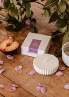 A white fluted soy wax Candle Refill - Artea by Mimi & August, a box labeled "Arte," half a plum, and pink petals rest on wood with leafy branches behind—a signature Mimi & August scene.