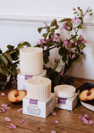 The Mimi & August Candle Refill - Artea, a ribbed white soy wax candle, is displayed on boxes with sliced plums and pink flowers atop a wooden surface, greenery in the background.