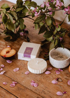 The Candle Refill - Artea by Mimi & August, displayed in a speckled ceramic cup with sliced plum, an Aries box, pink flower petals, and leafy branches on a wooden table, creates a serene and floral candle arrangement.