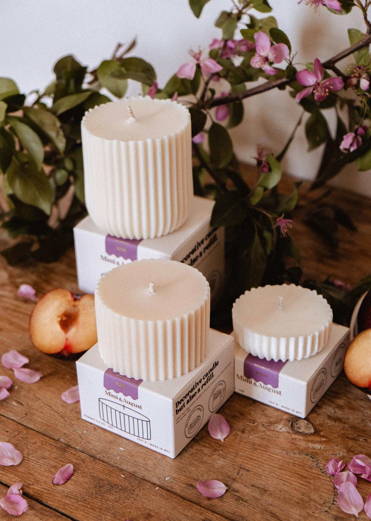 Three white ribbed Mimi & August Candle Refill - Artea candles rest on branded boxes atop wood, surrounded by peach slices, pink flower petals, and leafy branches—an ideal scene to showcase these floral soy candle refills.
