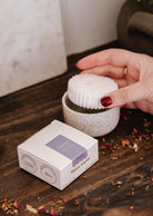 Une main soulève la Recharge de bougie Botanica de Mimi & August, en cire de soya blanche, d'un bougeoir en céramique moucheté; sa boîte et des pétales de fleurs séchées reposent sur une surface en bois.