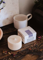 Une Recharge de bougie Botanica de Mimi & August, une boîte et une tasse blanche sont présentées sur une surface en bois avec des pétales de fleurs séchées éparpillés autour.