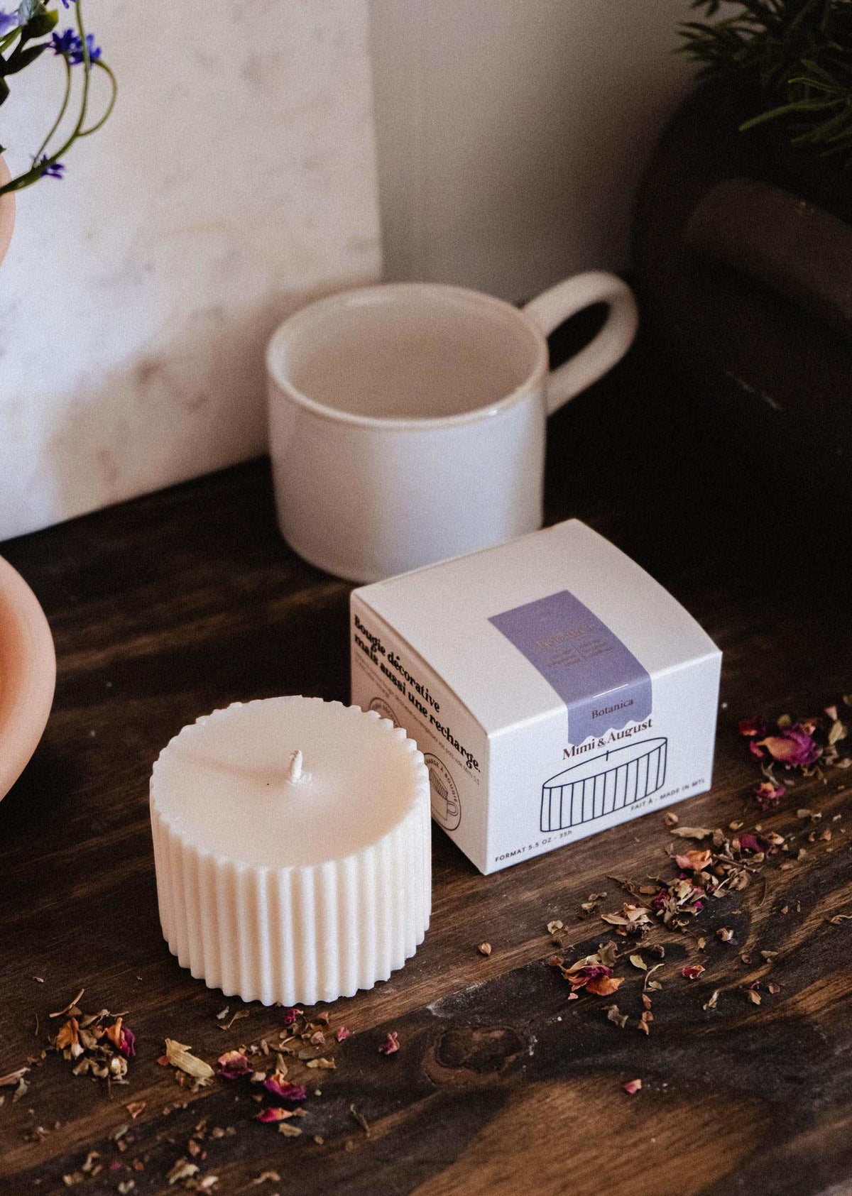 Une Recharge de bougie Botanica de Mimi & August, une boîte et une tasse blanche sont présentées sur une surface en bois avec des pétales de fleurs séchées éparpillés autour.