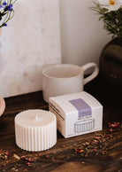 La Recharge de bougie Botanica de Mimi & August, une bougie de soya blanche et cannelée, repose sur une surface en bois à côté de sa boîte de marque, d'une tasse en céramique blanche et de pétales de fleurs séchées.