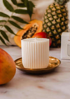 The Candle Refill Coco Mango by Mimi & August, a white ribbed tropical candle, sits on a gold plate with mango, papaya, pineapple, and green leaves in the background.