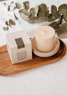 A ribbed, cream-colored eco-conscious candle in a ceramic dish sits on a wooden tray next to its box, with eucalyptus leaves behind—pair it perfectly with the Mimi & August Candle Refill Eucalipto.