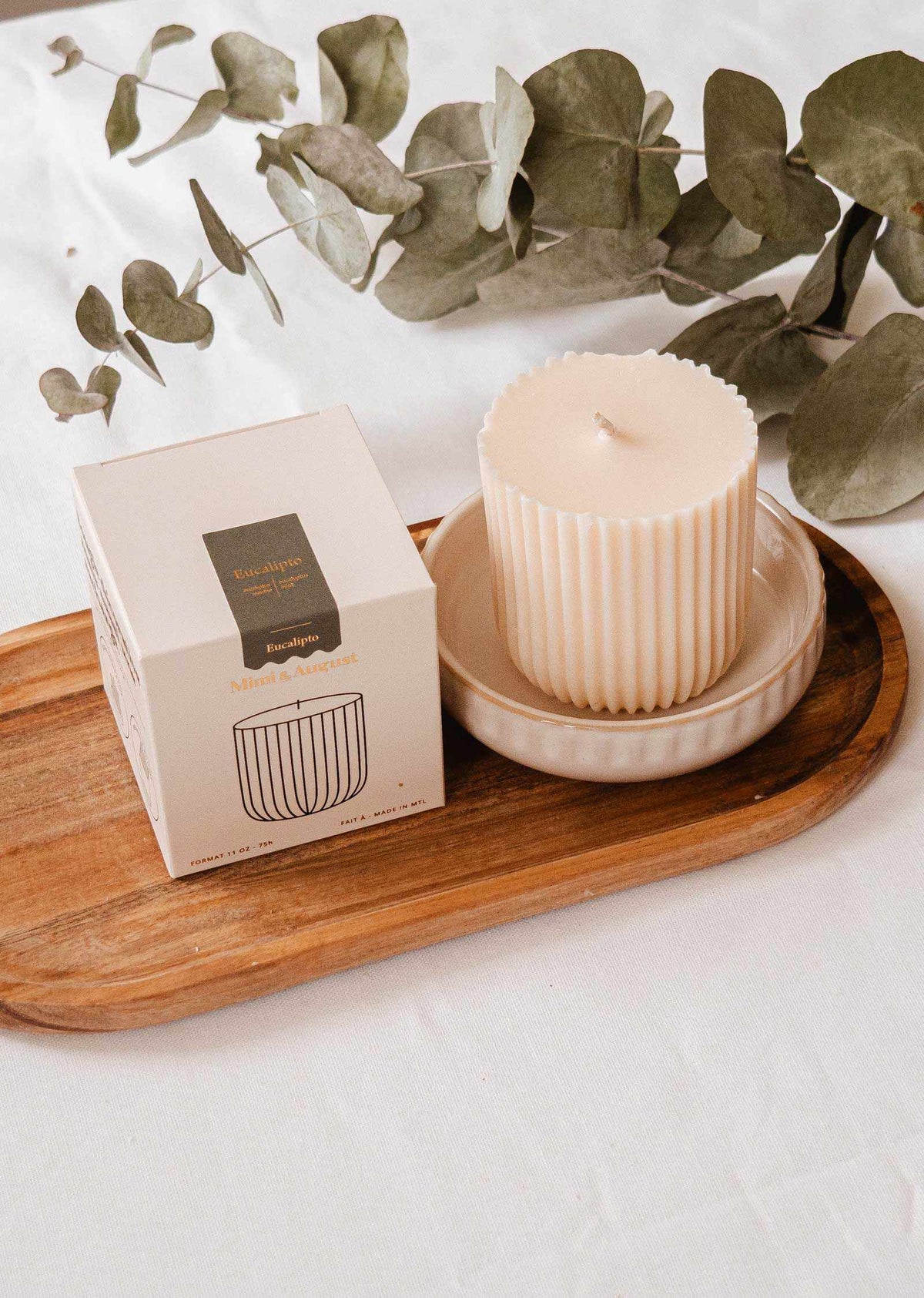 A ribbed, cream-colored eco-conscious candle in a ceramic dish sits on a wooden tray next to its box, with eucalyptus leaves behind—pair it perfectly with the Mimi & August Candle Refill Eucalipto.
