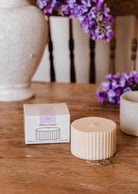 A ribbed cream-colored Candle Refill Lilac by Mimi & August sits next to its box on a wooden table, with a mug and purple flowers in the background.