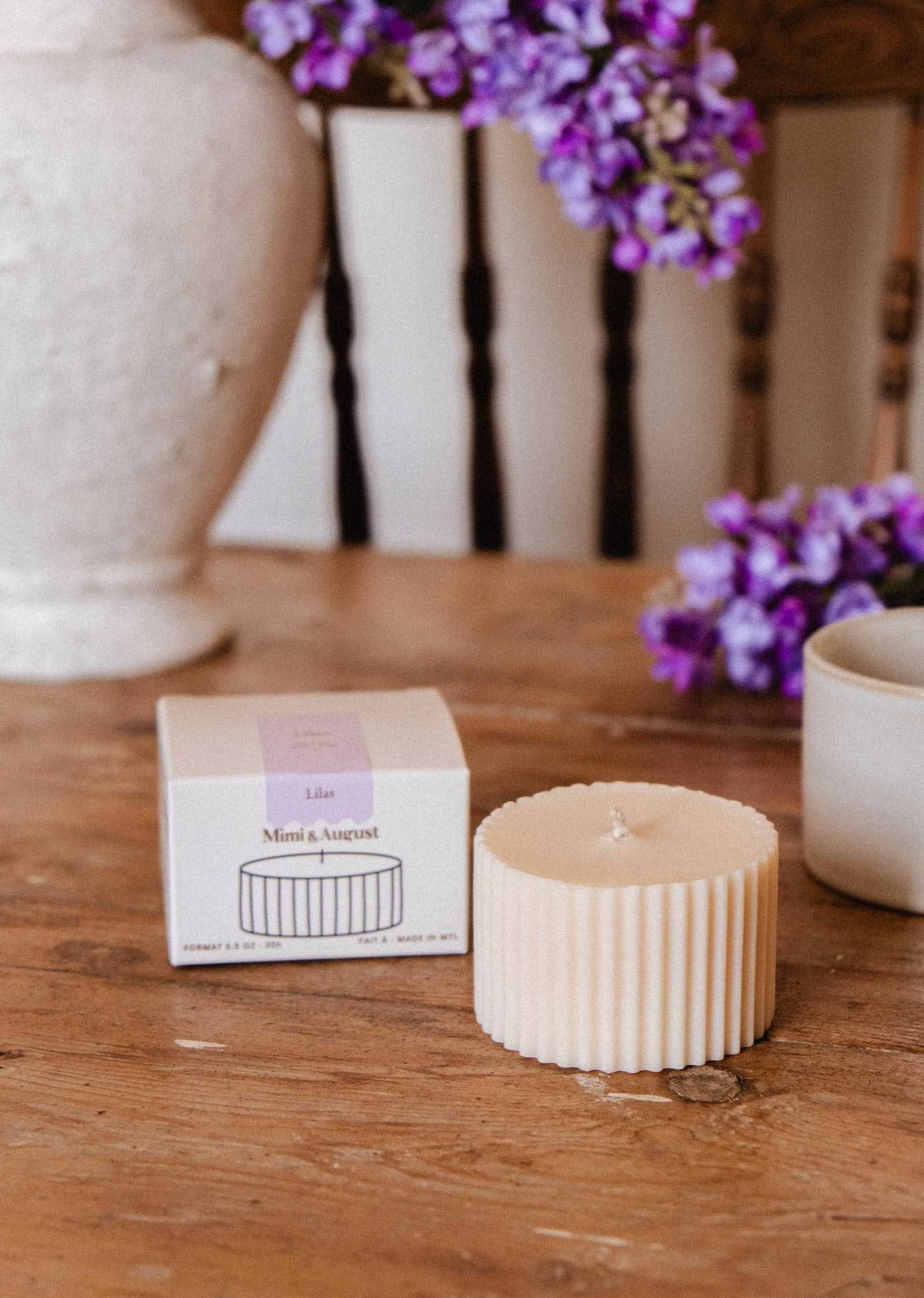 A ribbed cream-colored Candle Refill Lilac by Mimi & August sits next to its box on a wooden table, with a mug and purple flowers in the background.