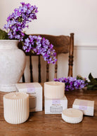 Four ribbed Mimi & August Candle Refill Lilac, crafted from natural soy wax in varying sizes, are displayed with their boxes on a wooden table beside a white vase of purple lilacs and a wooden chair in the background.