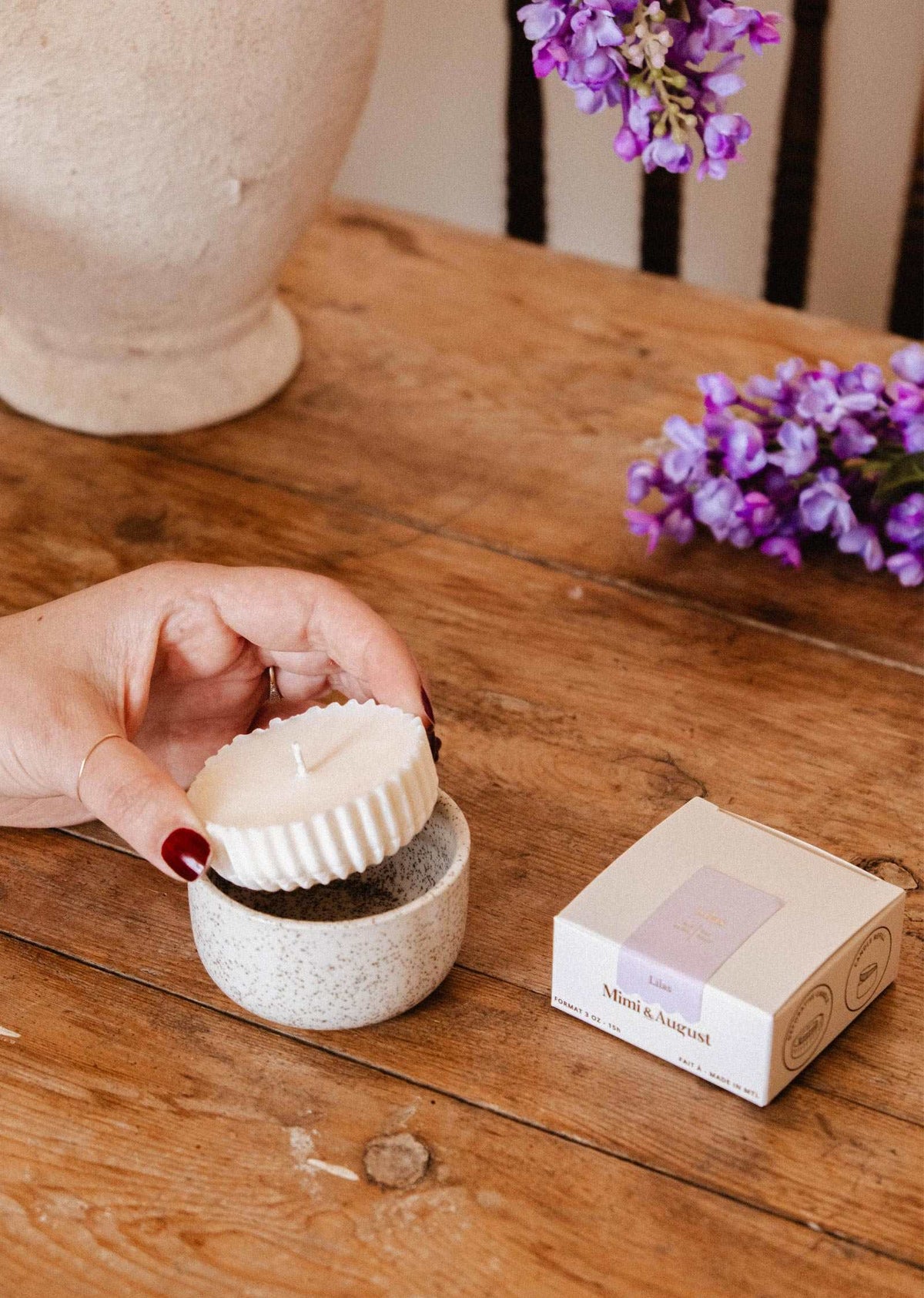 A hand places a Mimi & August Candle Refill Lilac, made of white ribbed soy wax, into a small ceramic container on a wooden table, with its box and purple flowers nearby, evoking the soothing scent of lilac.