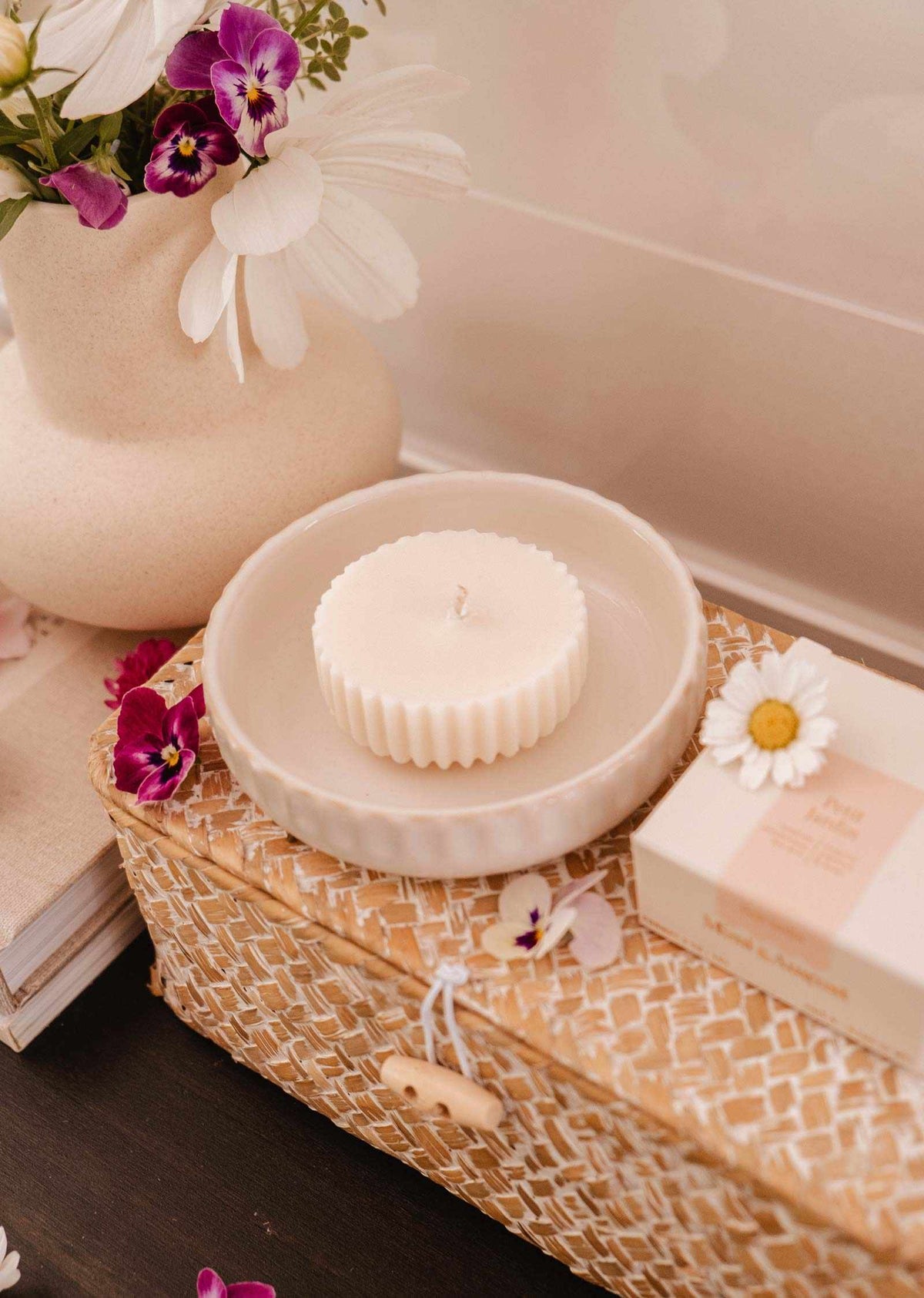 Une recharge de bougie ronde et blanche Petit Jardin de Mimi & August au parfum enchanteur repose dans un plat en céramique sur un panier tressé avec un fermoir bouton. Un vase blanc écoresponsable avec des fleurs violettes et blanches se trouve à proximité, accompagné d'une boîte posée sur des livres.