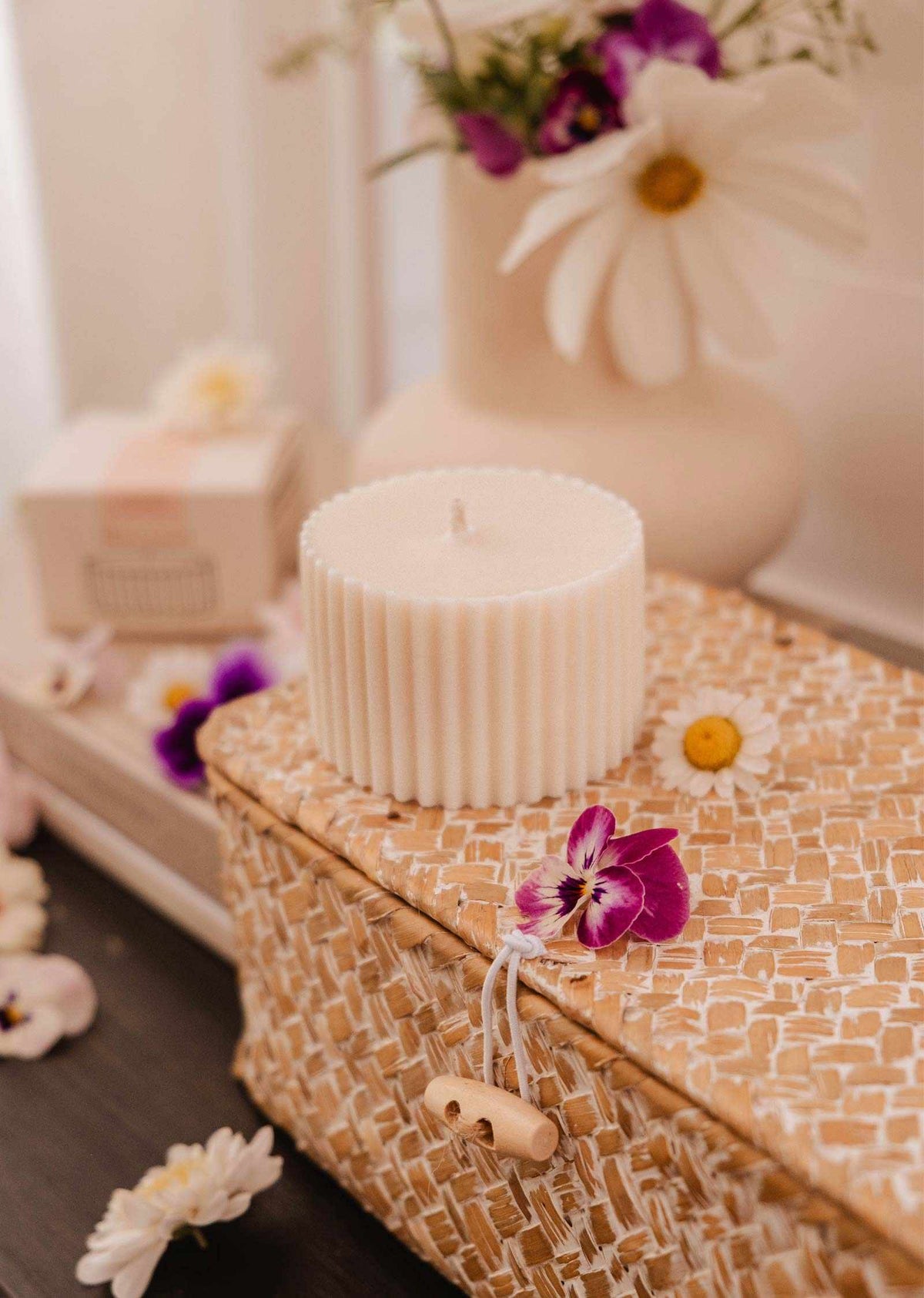 Une Recharge de bougie Petit Jardin de Mimi & August au parfum enchanteur repose sur un panier tressé, entourée de fleurs violettes et blanches. Un vase blanc avec des fleurs similaires en arrière-plan souligne l'ambiance écoresponsable.