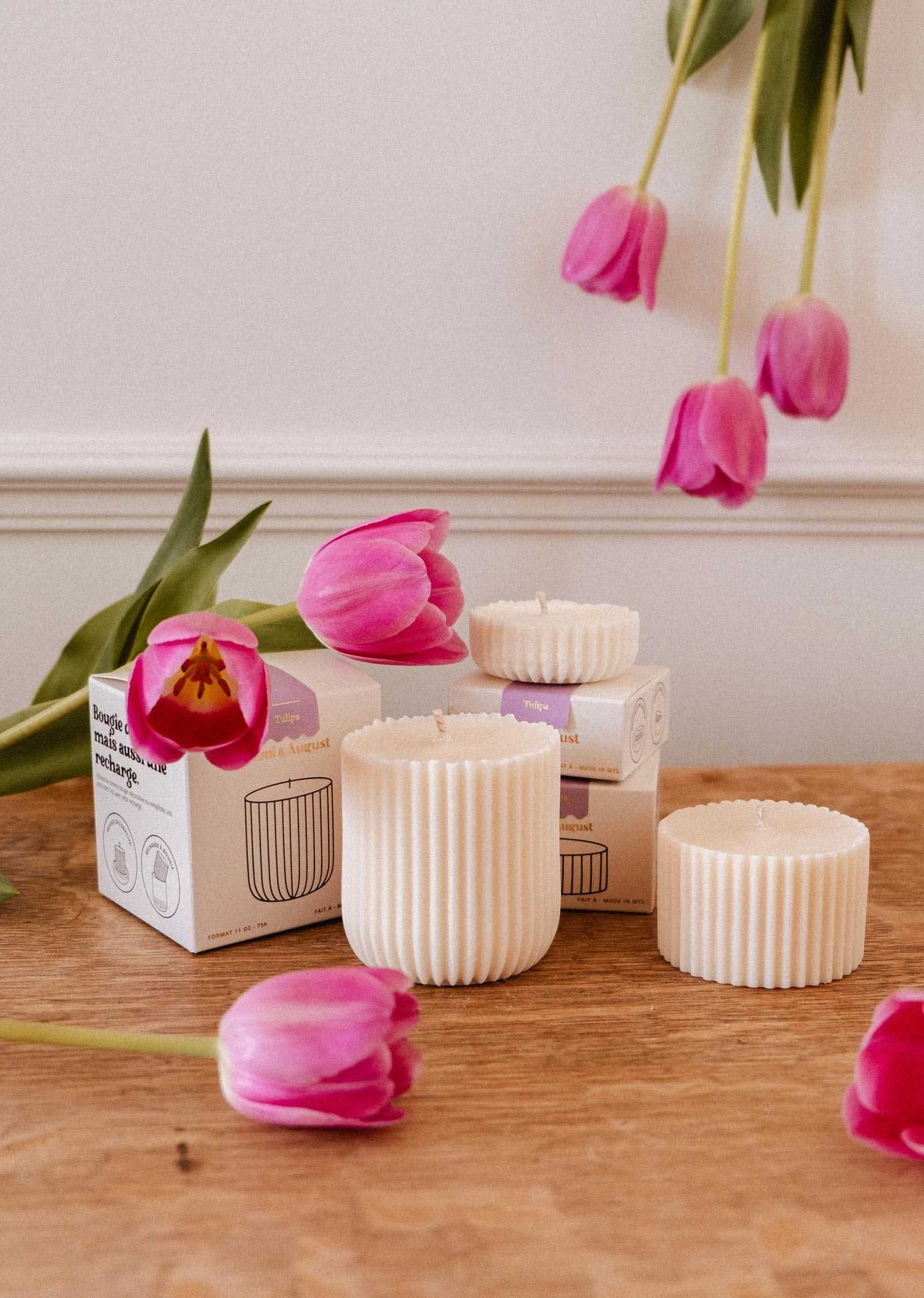 Three Mimi & August Candle Refill - Tulipa soy wax candles with ribbed white design sit in their boxes on a wooden surface, surrounded by pink tulips and set against a white wall.