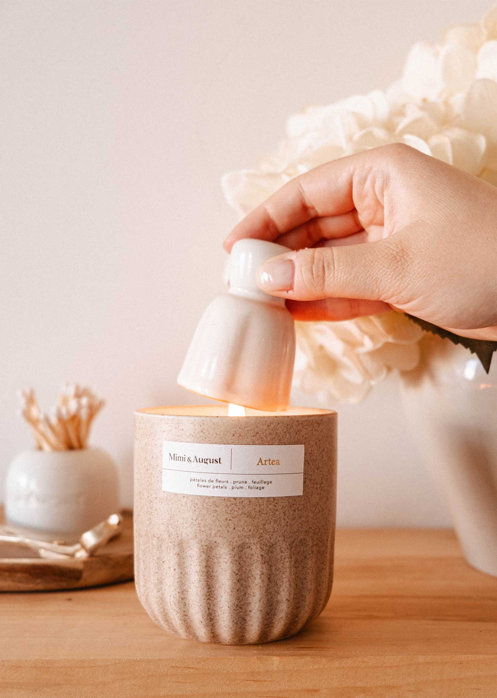 A hand uses the Mimi & August Candle Snuffer to extinguish a lit candle in a beige container on a wooden surface, with decorative candle accessories in the background.