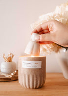 A hand uses the Mimi & August Candle Snuffer to extinguish a lit candle in a beige container on a wooden surface, with decorative candle accessories in the background.