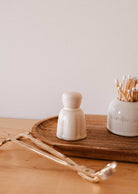 A Mimi & August Candle Snuffer rests on a light wood surface beside a small ceramic container, a jar of cotton swabs, and other candle accessories.