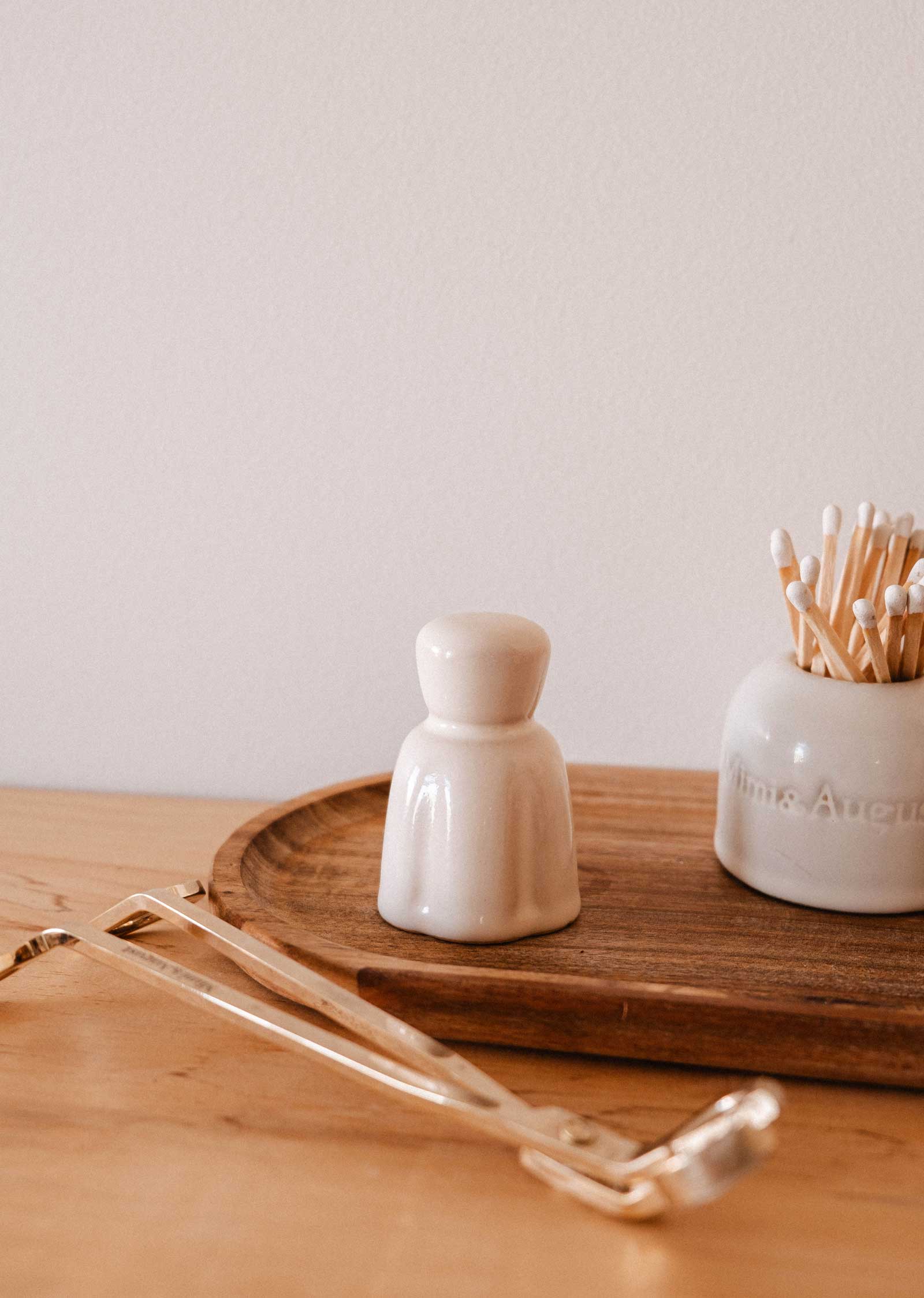 A Mimi & August Candle Snuffer rests on a light wood surface beside a small ceramic container, a jar of cotton swabs, and other candle accessories.
