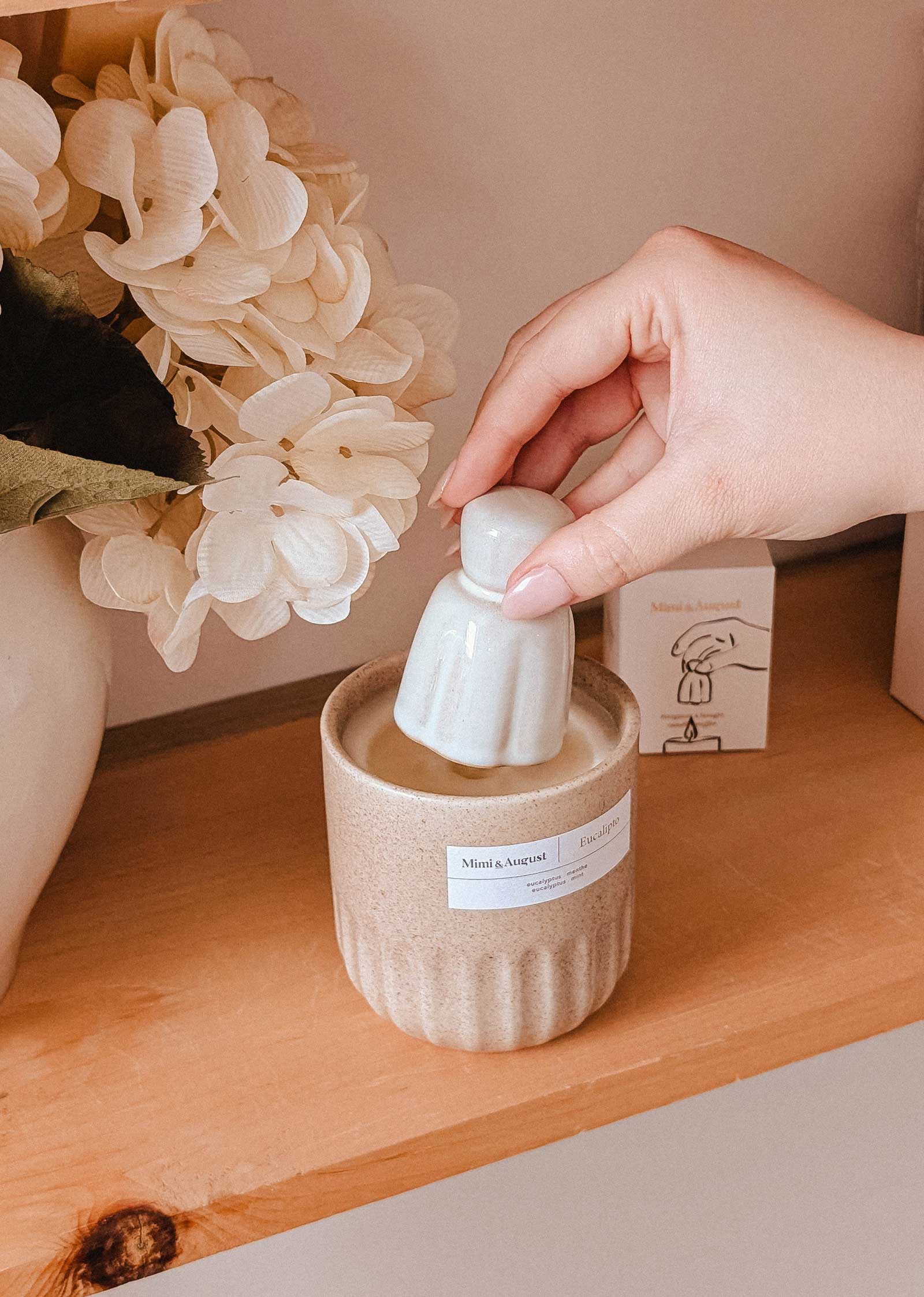 A hand sets the Mimi & August Candle Snuffer into a candle jar on a wooden shelf beside a vase of white flowers, bringing charm to essential candle accessories.