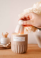 A hand uses the Mimi & August Candle Snuffer to extinguish a lit candle in a beige container on a wooden surface, with decorative candle accessories in the background.