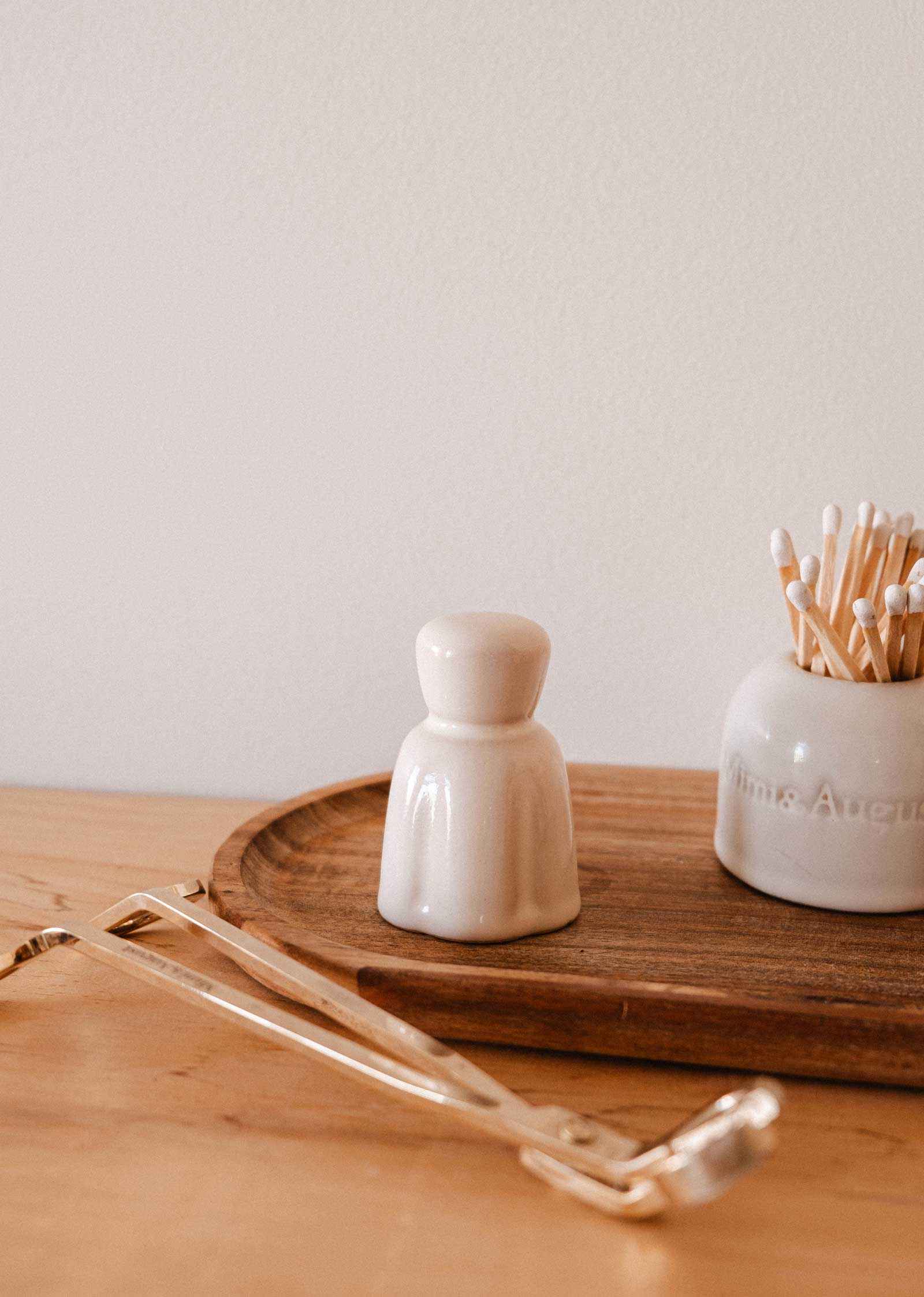 A Mimi & August Candle Snuffer rests on a light wood surface beside a small ceramic container, a jar of cotton swabs, and other candle accessories.