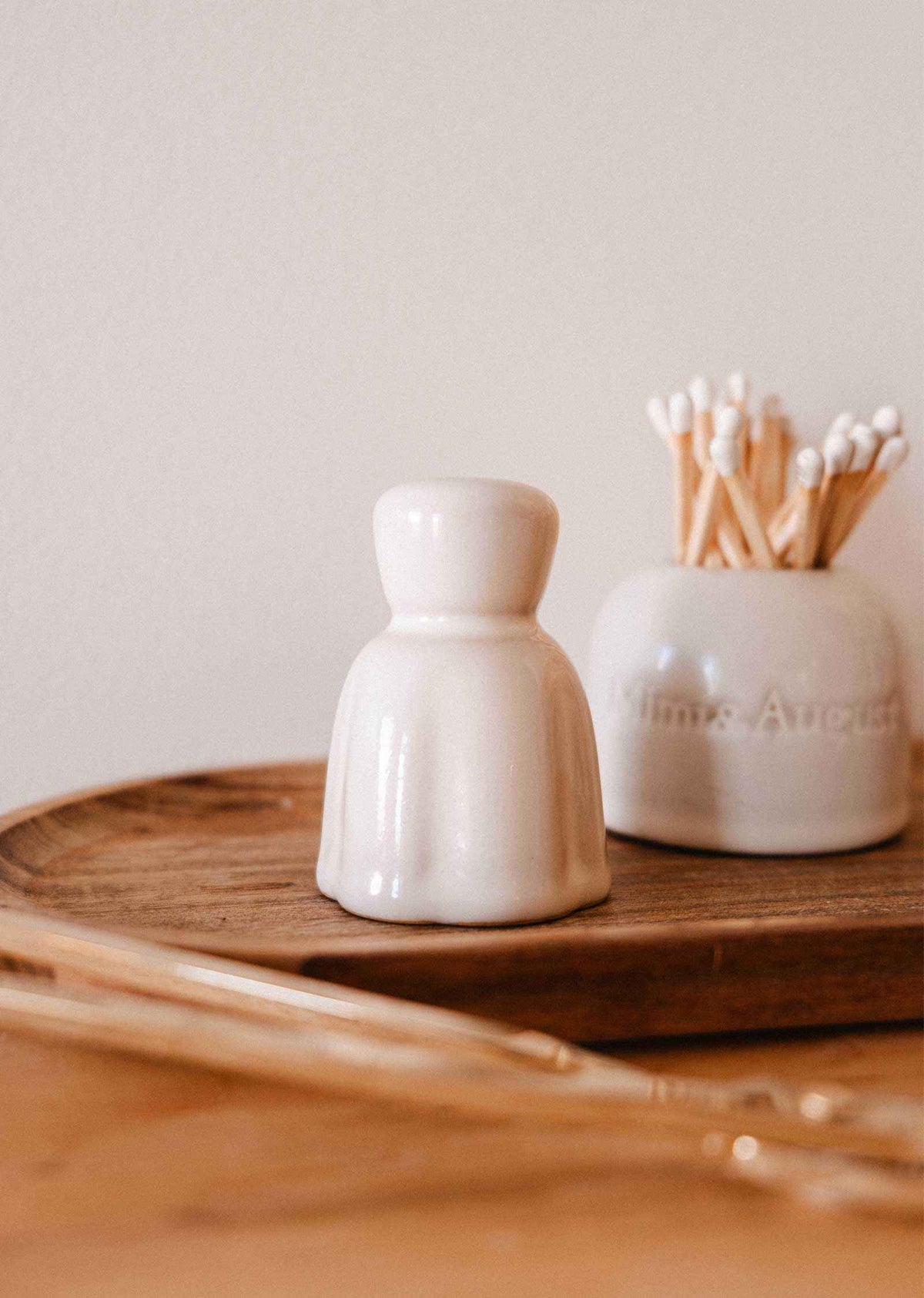The Mimi & August Candle Snuffer, a small white ceramic piece, sits on a wooden tray beside a jar of cotton swabs, adding an elegant touch to your candle accessories collection.