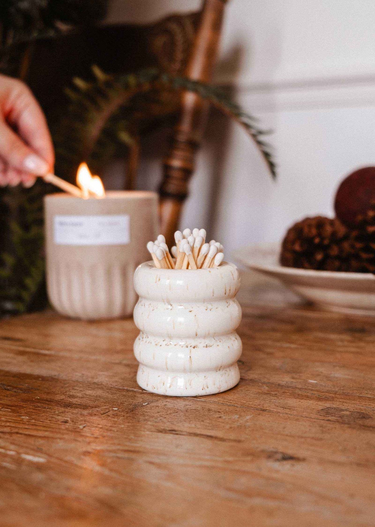 The Mimi & August Ceramic Match Pot, filled with wooden matches, sits on a wooden table as a hand lights a candle in the background, showcasing stylish candle accessories.