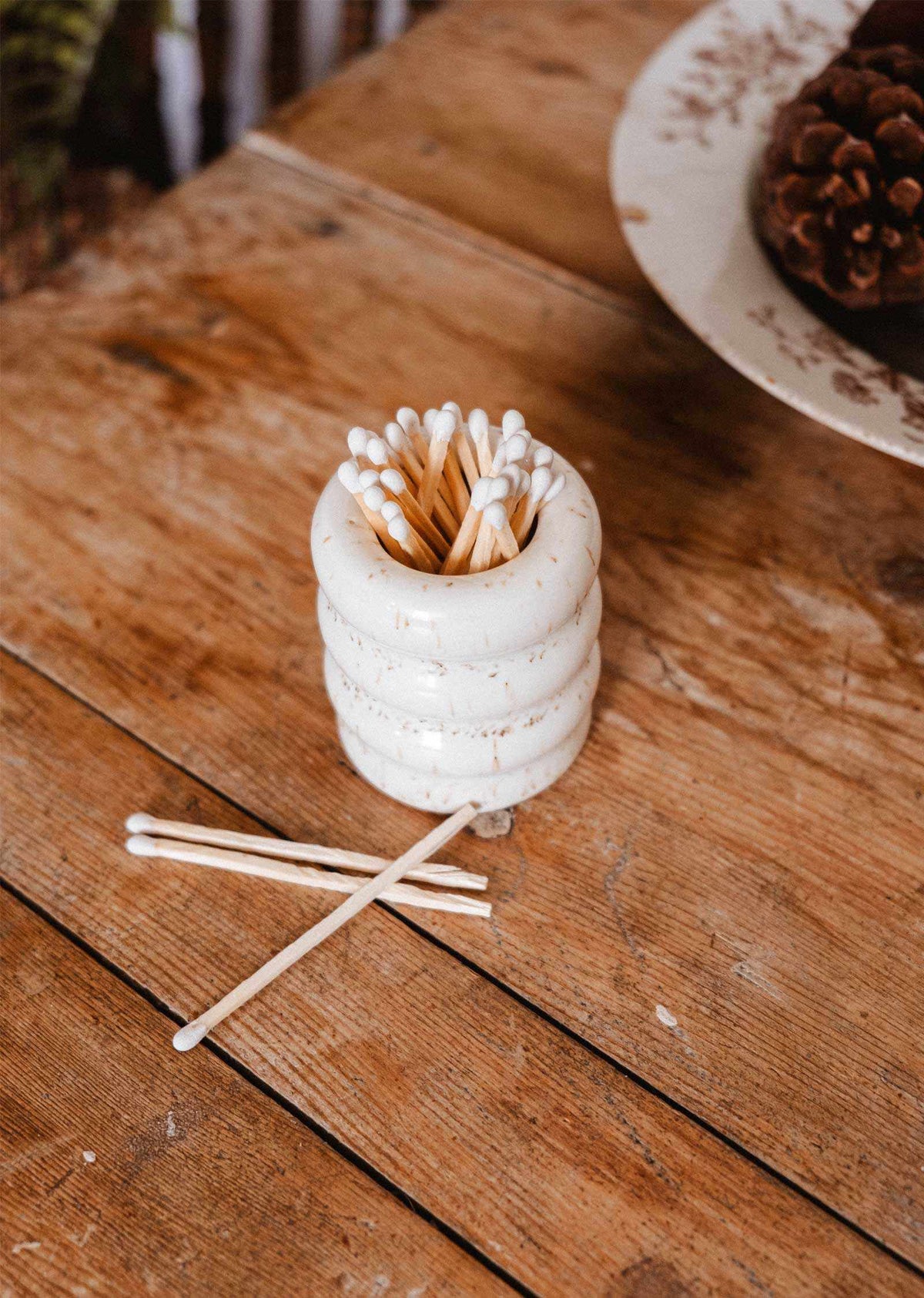 The Mimi & August Ceramic Match Pot, filled with cotton swabs, sits on a wooden table with four swabs beside it—perfect as a candle accessory for any space.