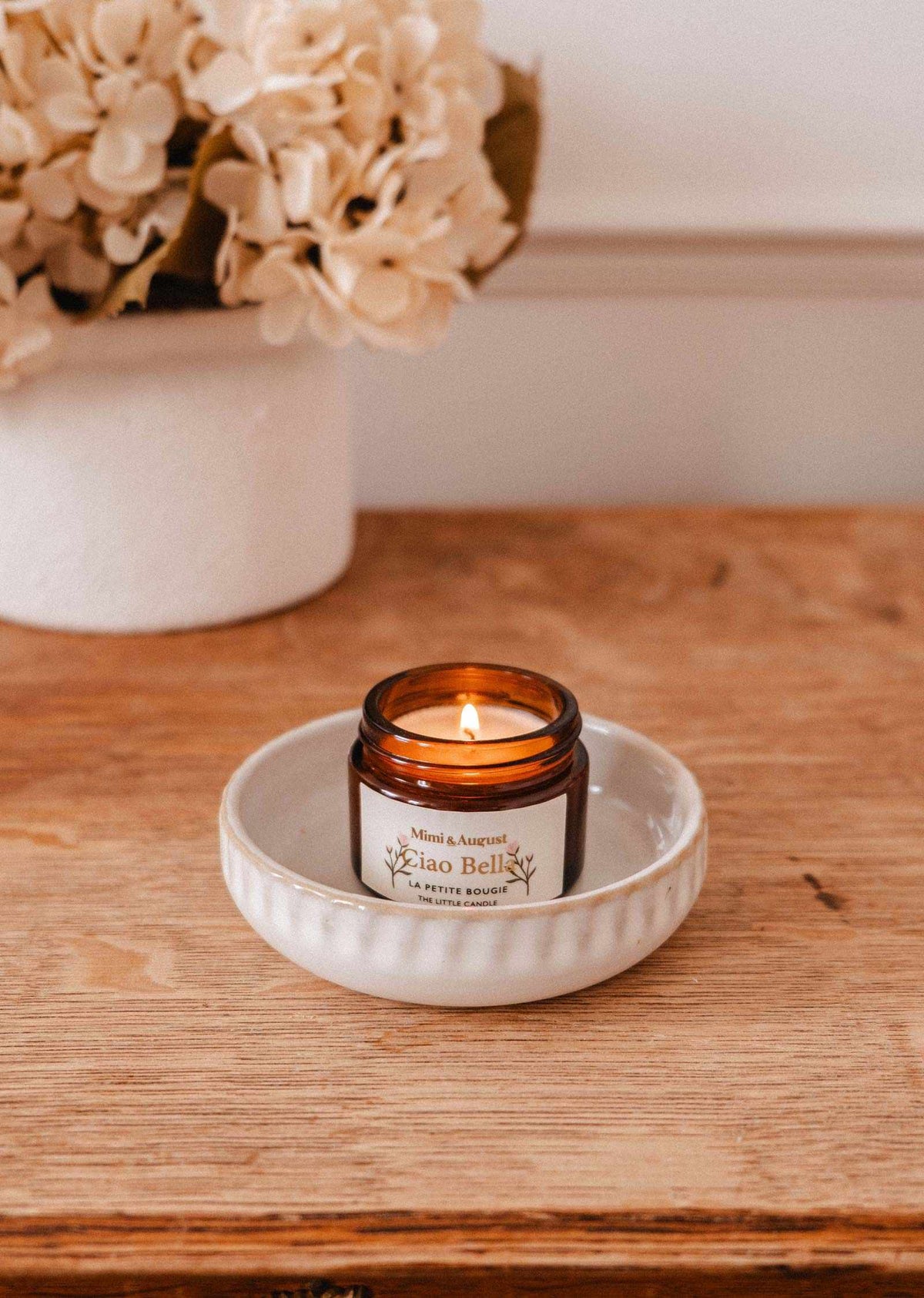 The Ciao Bella – 2 oz Soy Candle by Mimi & August, hand-poured in Montreal, rests in a brown glass jar on a wooden surface with cream-colored flowers in the background.