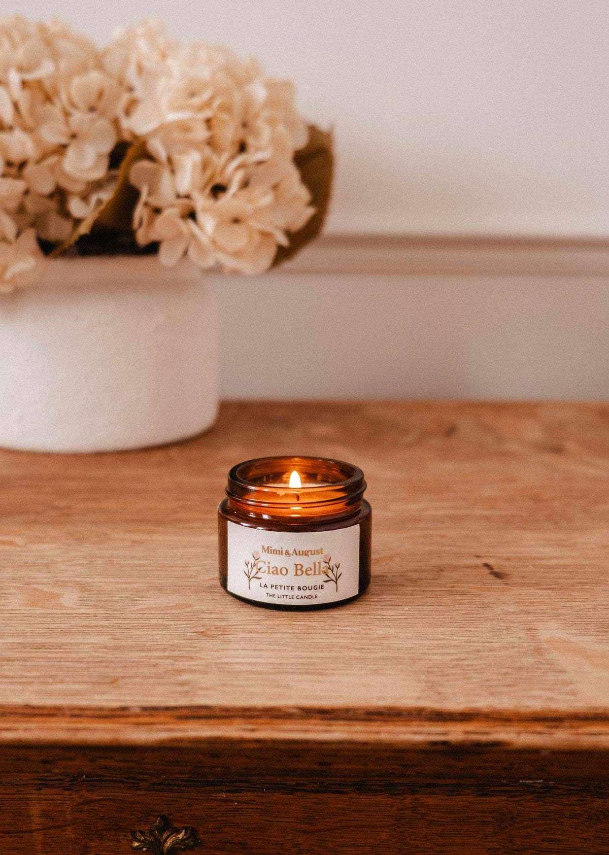 The Mimi & August Ciao Bella – 2 oz Soy Candle, hand-poured in Montreal, glows in a brown glass jar on wood beside a white vase with pale dried flowers.