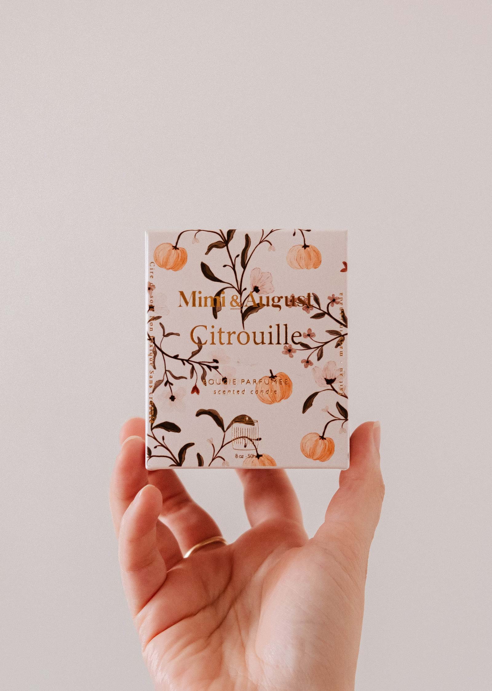 A hand holds a small "Citrouille - Reusable Candle" box from Mimi & August, decorated with orange pumpkins and branches, revealing a charming pumpkin spice candle inside.