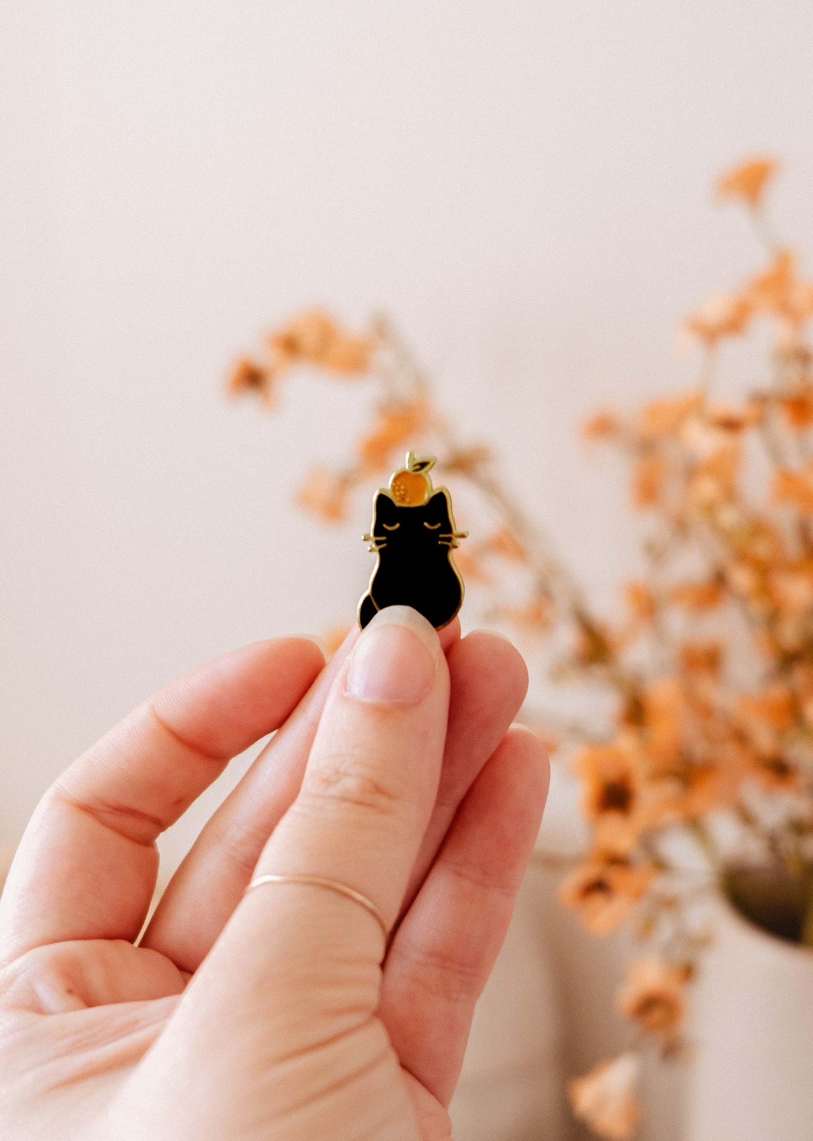 A hand holds the Clementine Cat Enamel Pin by Mimi & August, a quirky small black cat accessory, with a blurred background featuring orange dried flowers.