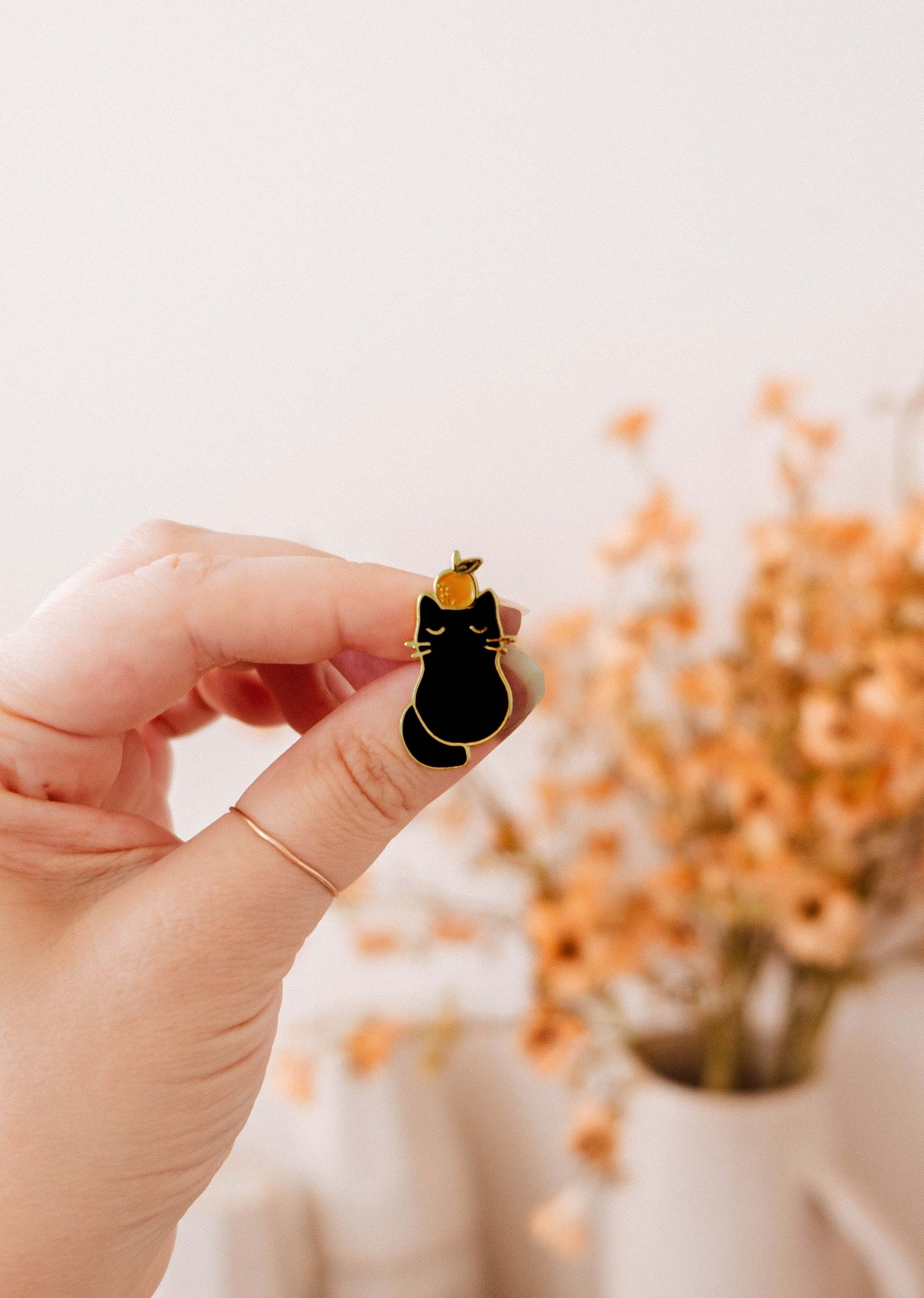 A hand holds the Mimi & August Clementine Cat Enamel Pin with gold outlines; a blurred vase of orange flowers appears in the background.