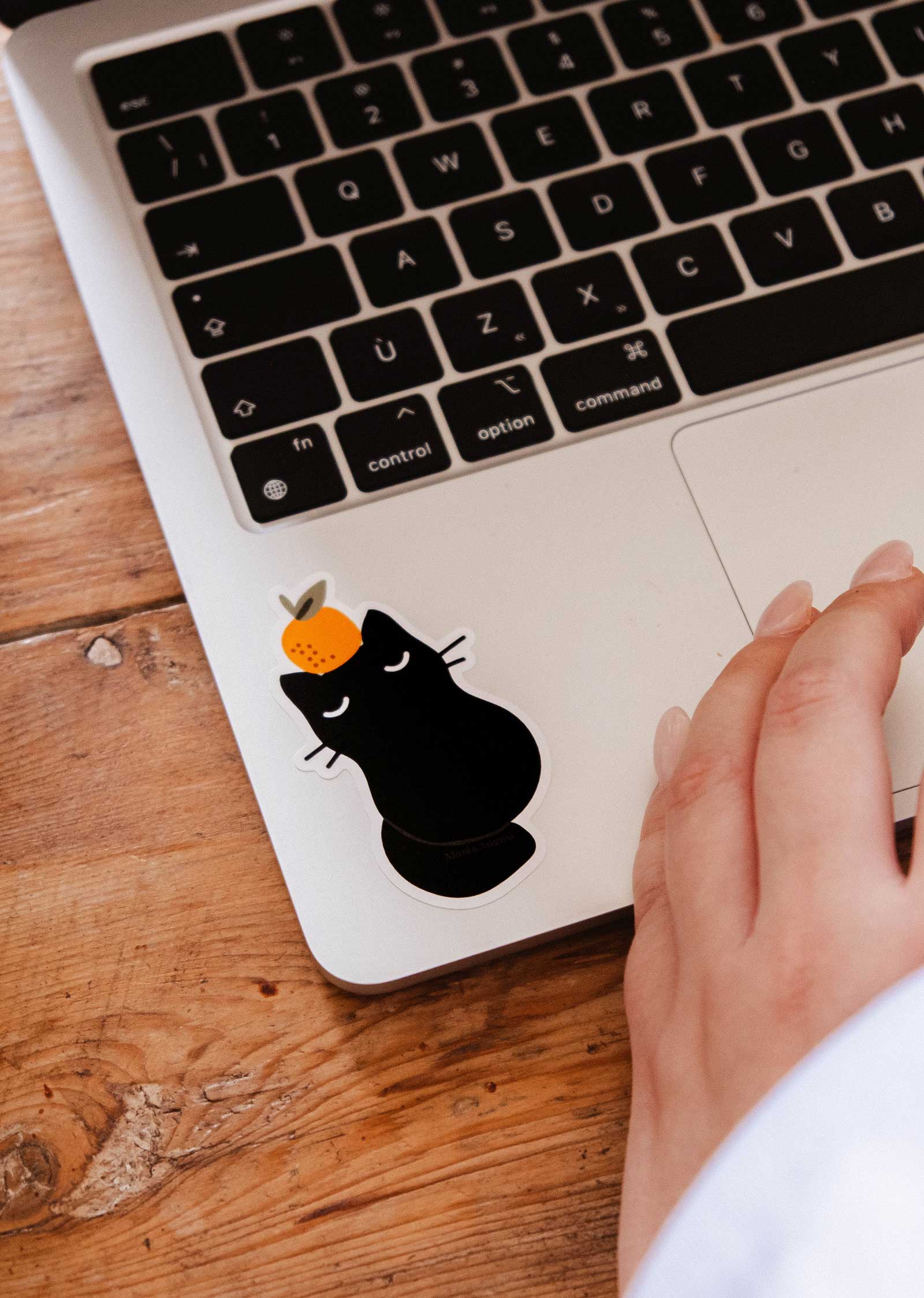 A hand rests on a laptop keyboard, which features Mimi & August's Clementine Cat - Vinyl Sticker—a black cat with an orange on its head—near the trackpad.