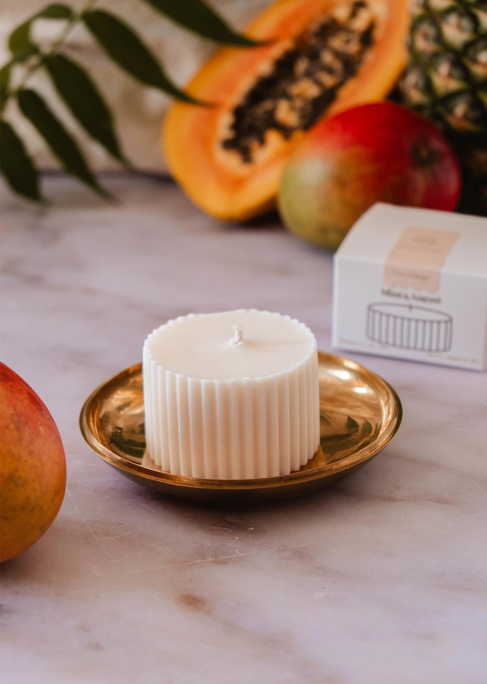 Une recharge de bougie - Coco Mango de Mimi & August placée sur une plaque en laiton est entourée de fruits tropicaux et d'une petite boîte blanche sur une surface en marbre.