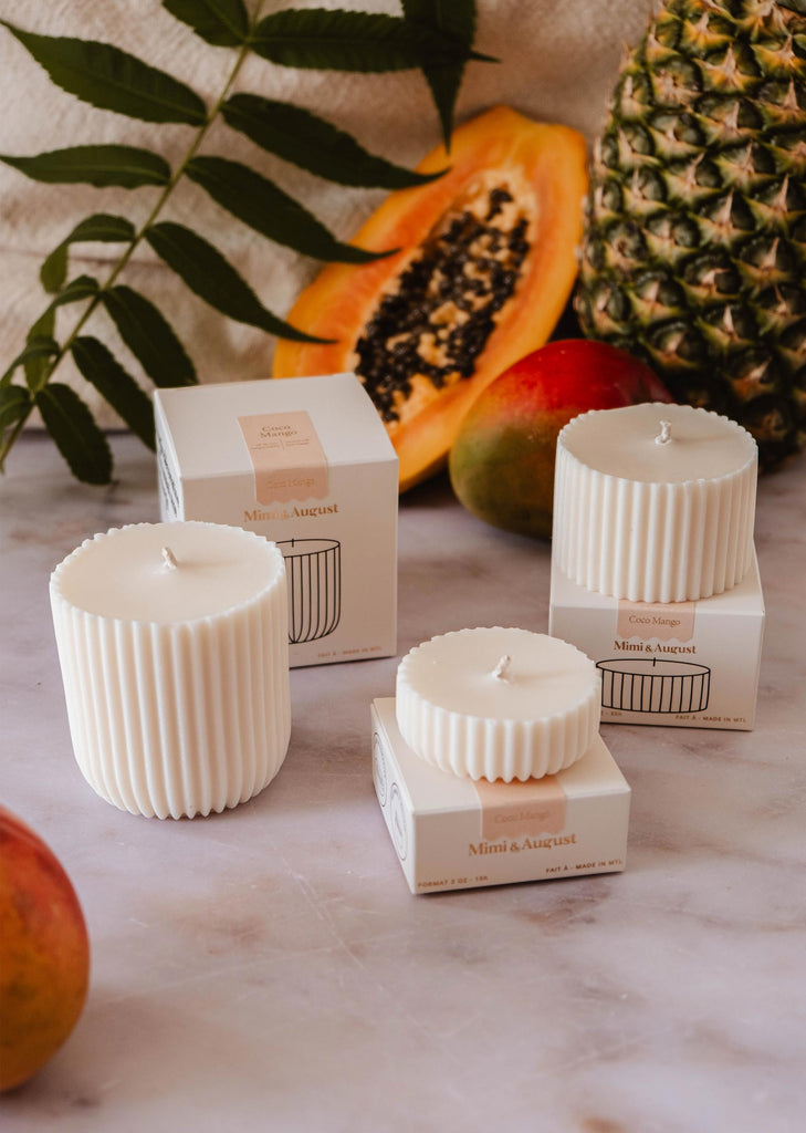 Three white ribbed candles on a marble surface, each placed near a matching box. Surrounding the candles are tropical fruits like papaya, pineapple, and mango, along with green foliage. Enjoy the ambiance with our eco-friendly Candle Refill - Coco Mango from Mimi & August for an exotic touch.