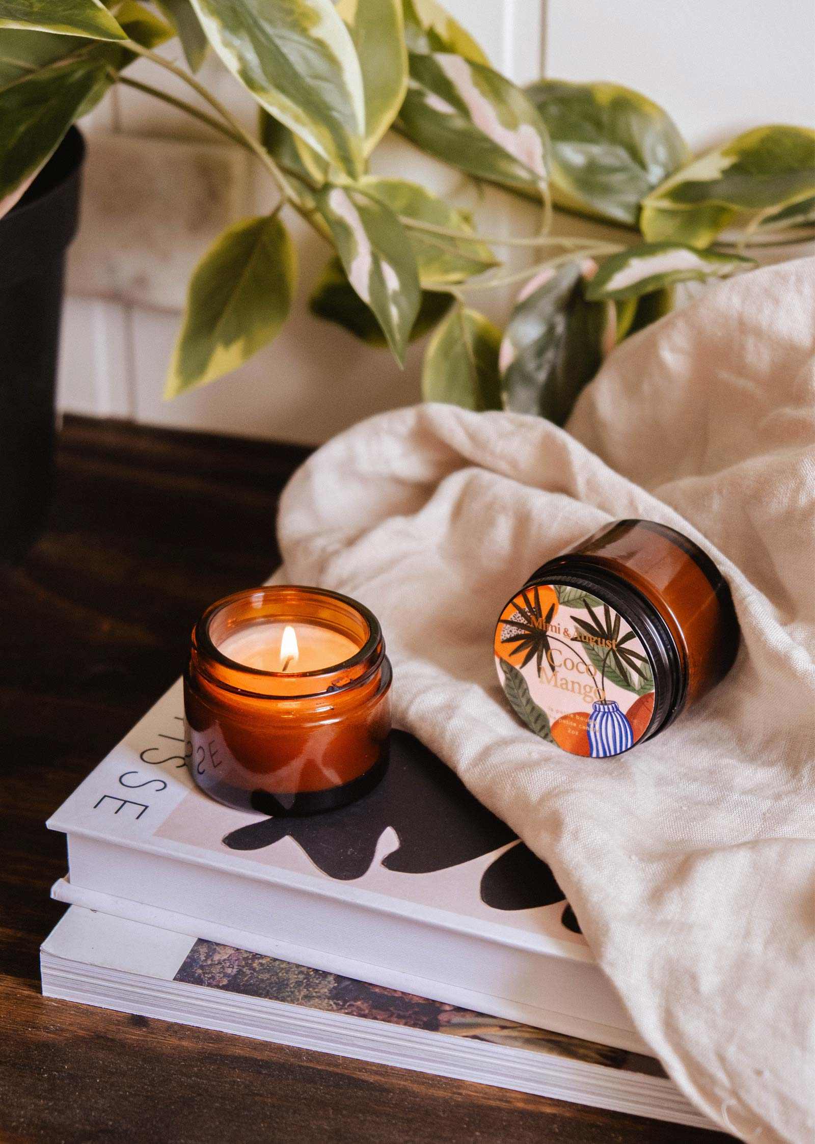 A lit Mimi & August Coco Mango - 2 oz candle fills the air with tropical fruit aroma, while another reusable jar rests on stacked books near a beige cloth and leafy plant atop a wooden surface.