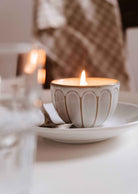 A lit Le Petit Café Mimi – Two Little Cups Candle in a fluted ceramic cup sitting on a matching saucer.