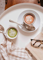 Le Petit Café Mimi – Two Little Cups Candle Set with green matcha and coffee designs sit on a white table next to a book and glasses.
