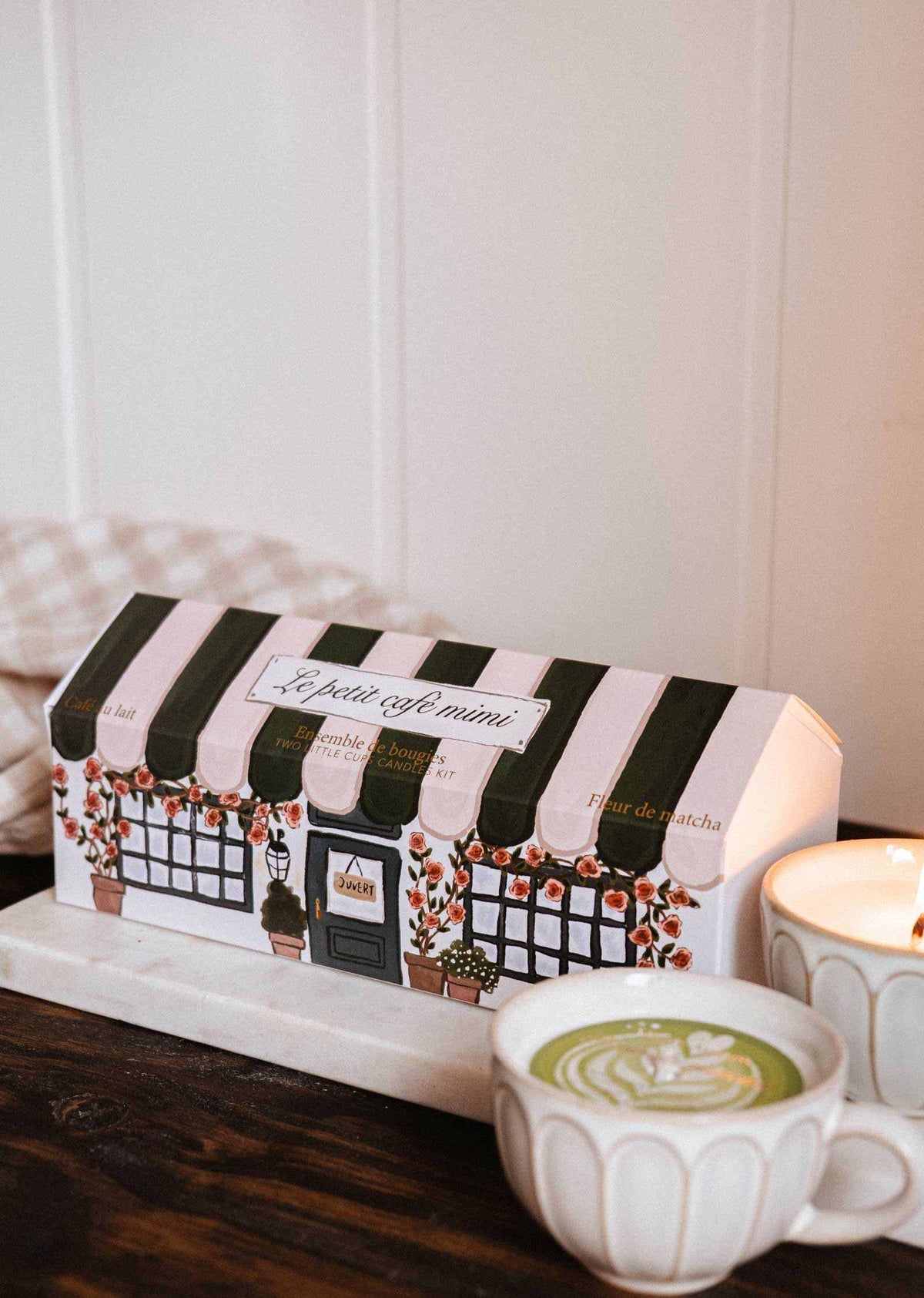 A Le Petit Café Mimi – Two Little Cups Candle Set designed to look like a cafe sits next to two lit soy candles in scalloped cups on a wooden surface.
