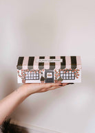 A person holds a Le Petit Café Mimi – Two Little Cups Candle Set, designed as a small café with black and white striped awnings and floral window boxes.