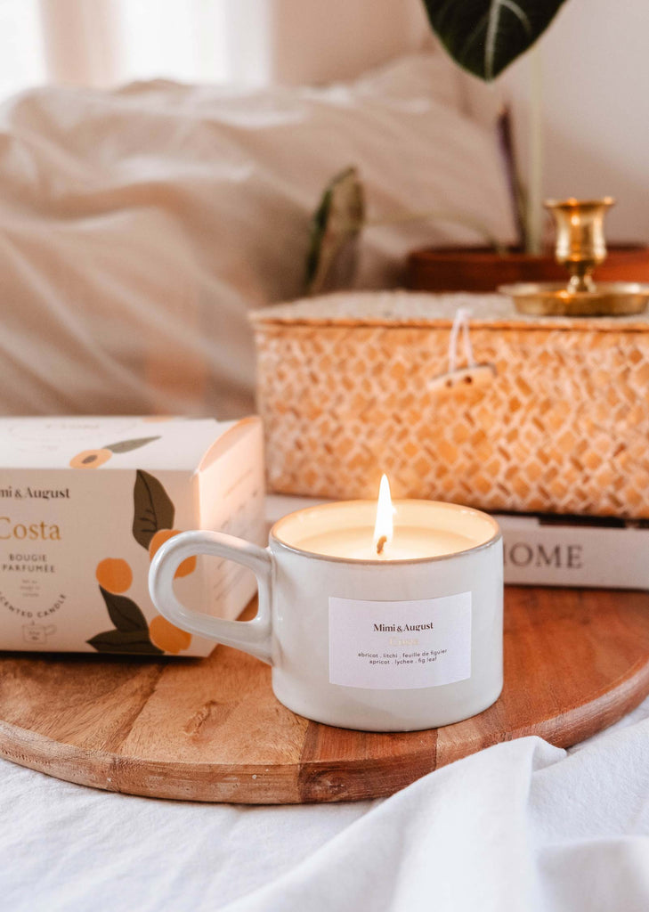 A bougie de soja Costa de Mimi & August à l'abricot et à la figue fraîche dans un mug blanc sur un plateau en bois, à côté d'un livre fermé et d'une plante décorative, sur un fond doux et confortable.