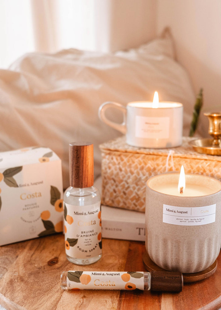 A cozy bedroom corner with lit Costa reusable candles from Mimi & August, essential oils, and a bed with white linens, creating a serene and inviting atmosphere.