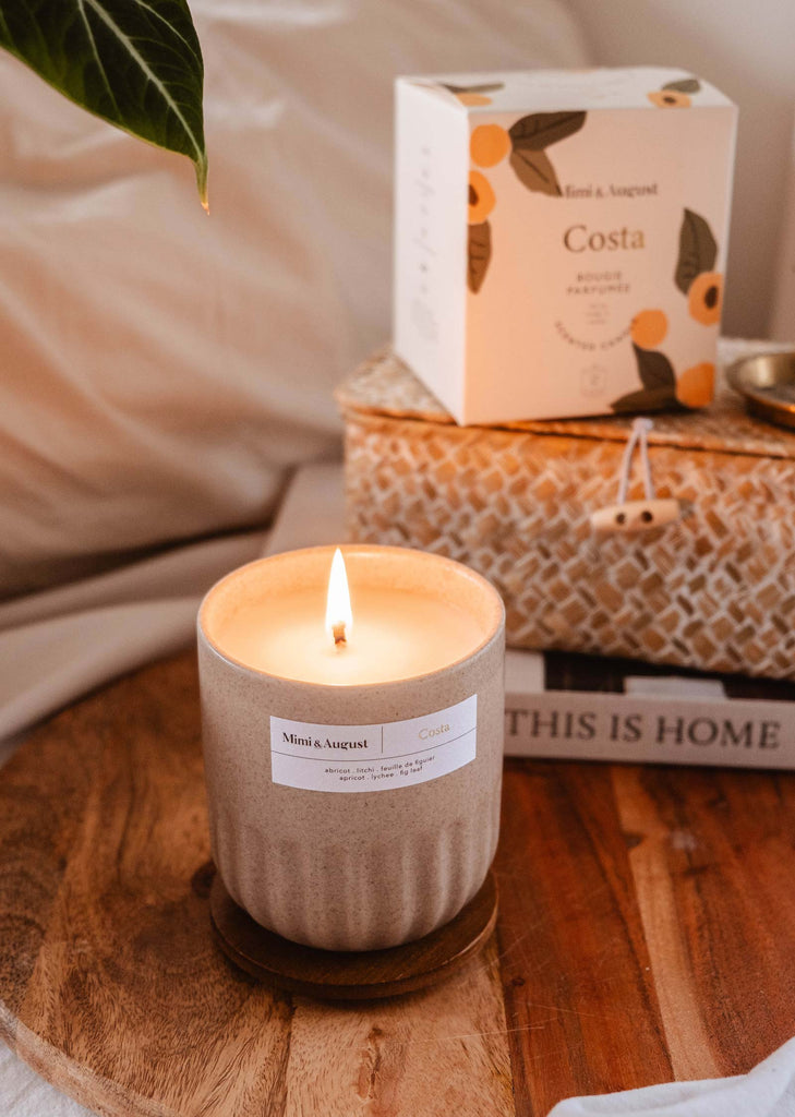 Une bougie réutilisable Costa dans un contenant beige avec une étiquette de parfum "abricot et figue fraîche", à côté d'une plante et d'une boîte portant la même marque Mimi & August, sur une surface en bois.