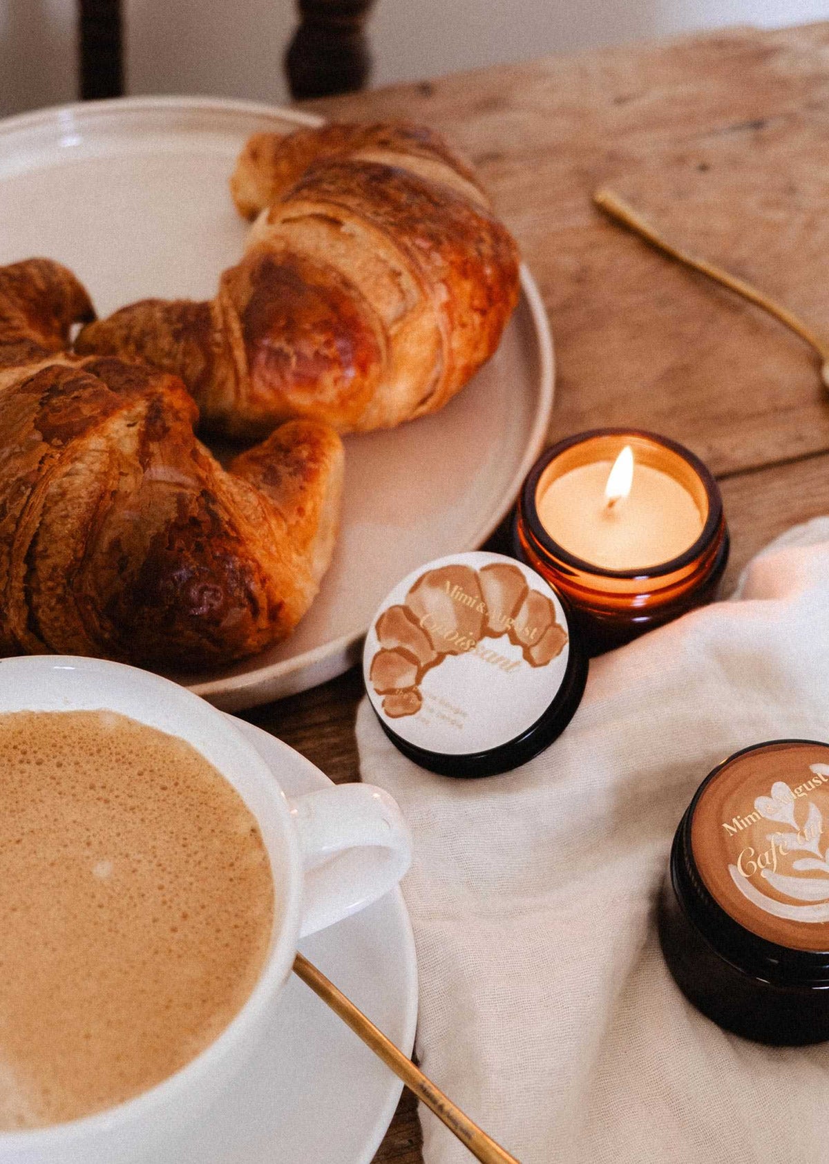 A Mimi & August Croissant – 2 oz Soy Candle sits on a wooden table with a white cloth, its subtle French bakery scent filling the air beside two croissants, a cup of coffee, and two skincare cream jars.
