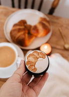 A hand holds Mimi & August’s Croissant – 2 oz Soy Candle in front of a table with a pastry, coffee, and a lit soy candle—invoking the cozy scent of a French bakery.